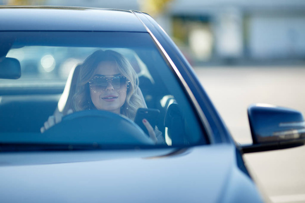 Image of beautiful blonde in sunglasses sitting in black car during day. - Φωτογραφία, εικόνα