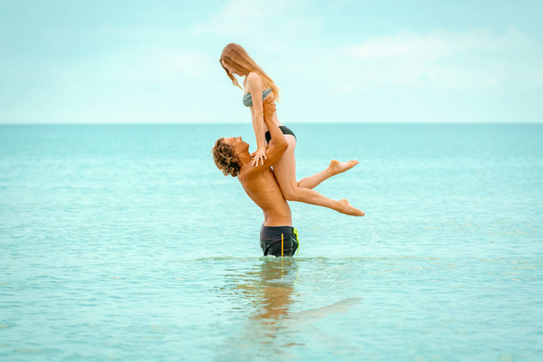 Woman jumps to the man in his arms, standing in the sea. Both are in the swimsuits. Smiling playful young couple in love having fun at sandy beach. - Photo, Image