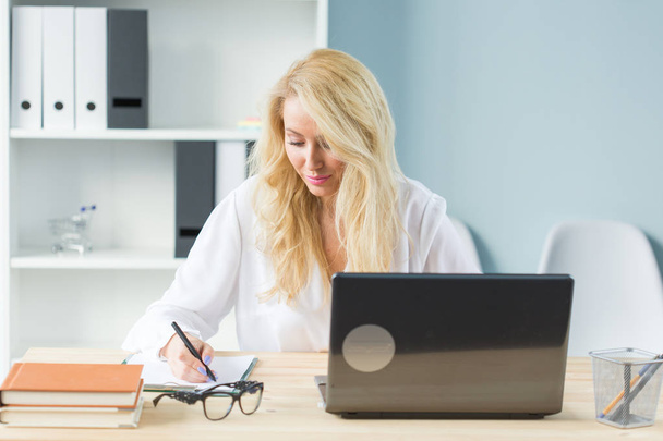 Τεχνολογία, εργασία και άτομα έννοια - όμορφη ξανθιά γυναίκα κάθεται στο γραφείο της και να εργάζονται στο lap-top - Φωτογραφία, εικόνα