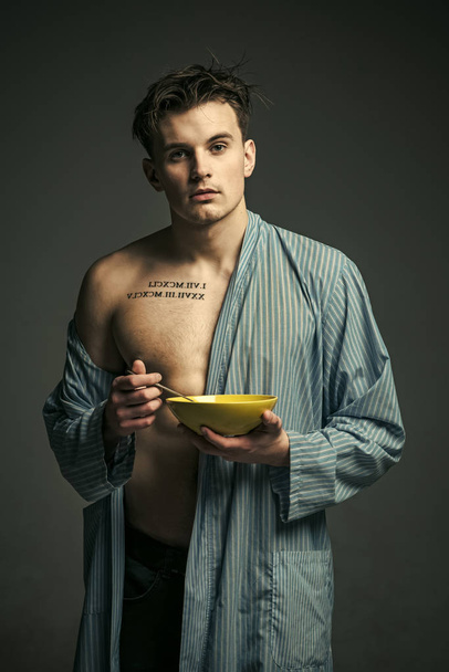 Guy in bathrobe with plate and spoon in hands. Man on calm face, muscular figure, holds plate with healthy food. Sporty nutrition concept. Man with muscular torso, looks attractive, dark background. - Foto, afbeelding