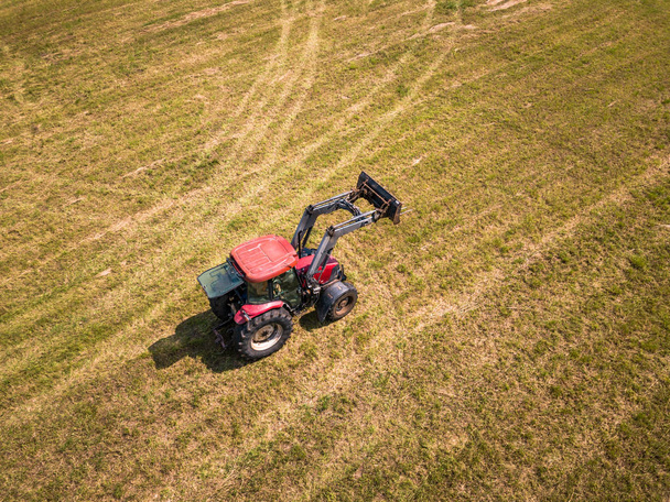 Εναέριο κηφήνα φωτογραφία γεωργός συγκομιδής σανού ρολά στο πεδίο σιτάρι με ένα κόκκινο τρακτέρ - ηλιόλουστη μέρα του καλοκαιριού - Φωτογραφία, εικόνα