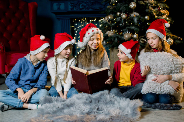 Beautiful little children read a book with Christmas stories on the background of a Christmas tree in red New Year hats - Photo, image