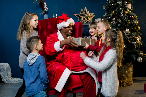 African Santa Claus sits on a background of a Christmas tree and presents gifts to the children - Фото, изображение