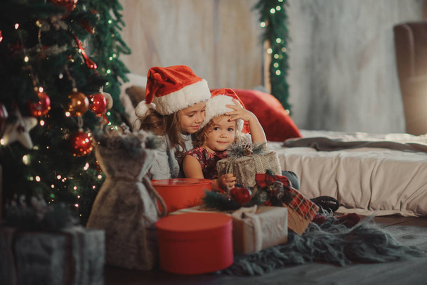 メリー クリスマスとハッピー ホリデー!2 つのかわいい小さな子供女の子姉妹は、クリスマスの魔法のギフト ボックスを開きます。正月飾りとクリスマス ツリーと居心地の良い部屋  - 写真・画像