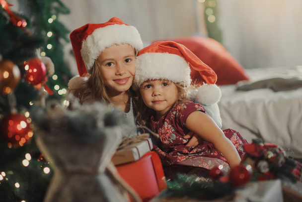 Merry Christmas and Happy Holiday! Two cute little children girls sisters open magic gift box on Christmas. Cozy  room with New year decoration and Christmas tree  - Photo, image