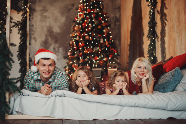 Merry Christmas and Happy Holidays! Cheerful parents and  and two little children having fun and playing together near Christmas tree on the bed in living room - Fotografie, Obrázek