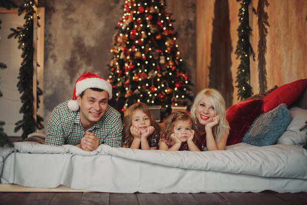 З Різдвом та Різдвом Христовим! Веселі батьки і двоє маленьких дітей розважаються і граються разом біля ялинки на ліжку у вітальні
 - Фото, зображення