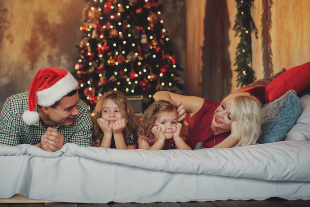 Merry Christmas and Happy Holidays! Cheerful parents and  and two little children having fun and playing together near Christmas tree on the bed in living room - Photo, Image