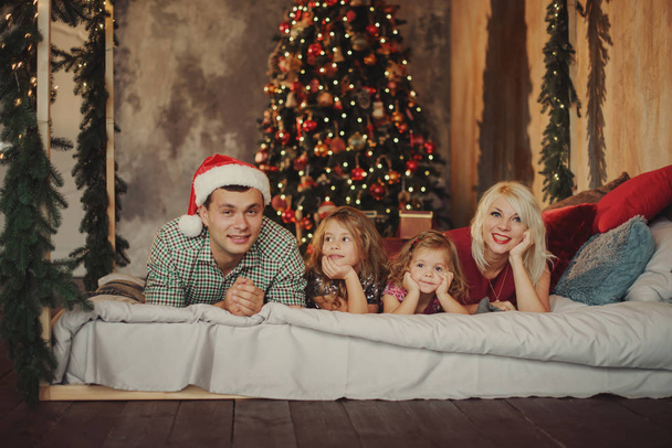 Merry Christmas and Happy Holidays! Cheerful parents and  and two little children having fun and playing together near Christmas tree on the bed in living room - Photo, Image