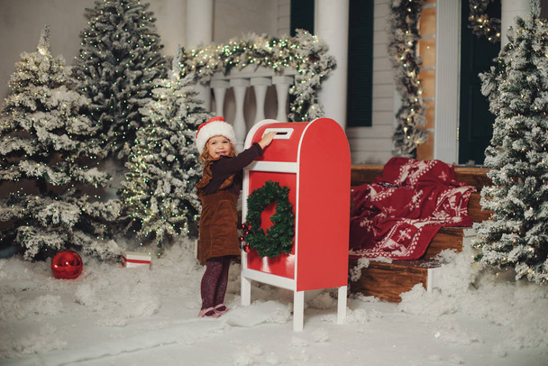 A little baby girl in a white knitted hat makes a wish and writes a letter to Santa Claus and sends a postcard by mail. Throws envelope in the mailbox - Foto, Bild
