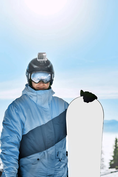Snowboarder con cámara de acción en un casco. Gafas de esquí con el reflejo de montañas nevadas. Retrato del hombre en la estación de esquí en el fondo cielo azul, mantenga snowboard. Usar gafas de esquí
 - Foto, Imagen