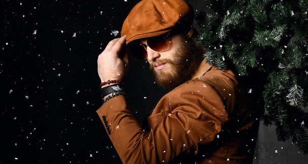Bearded man in brown leather jacket and cap hold Christmas tree under snow - Φωτογραφία, εικόνα