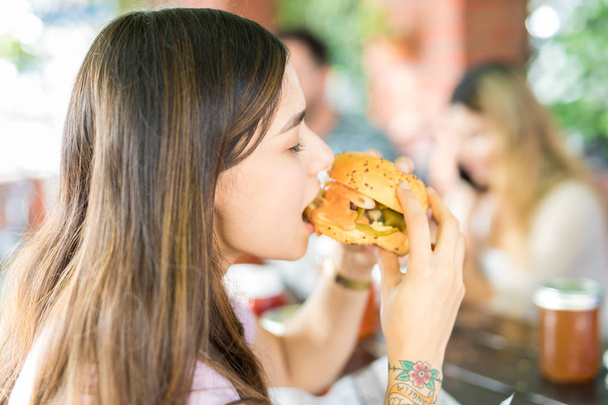 Vista lateral de la linda mujer joven comiendo deliciosa hamburguesa con queso en el restaurante al aire libre
 - Foto, imagen
