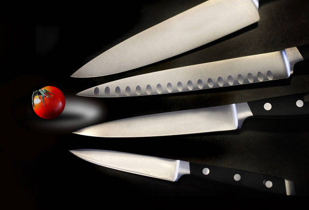 Assorted sharp and shiny kitchen knives are seen with a small tomato in this photograph. - Foto, Imagem