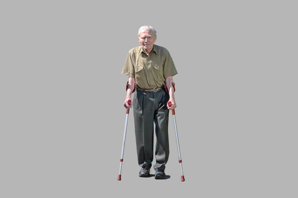 Hombre jubilado mayor caminando en muletas en una pasarela entre setos verdes en un suburbio residencial acercándose a la cámara frontal en
 - Foto, Imagen