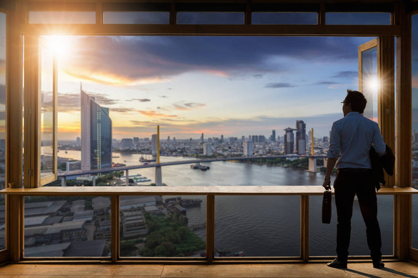Concept vision, Young businessman wearing comfortable casual suit jacket standing in working room near the glass window and looking through the modern buildings in the skyline. - Photo, Image