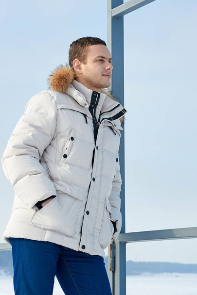 Fashion portrait of the young beautiful man in a white jacket with fur hood in winter - Photo, image