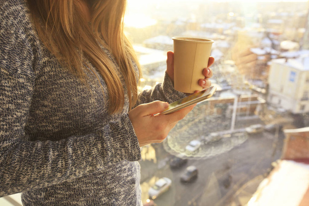 Nuori nainen pitää kuppi kahvia ja matkapuhelinta käsissä ja seisoo lähellä ikkunaa kaupunkimaisema tausta
 - Valokuva, kuva