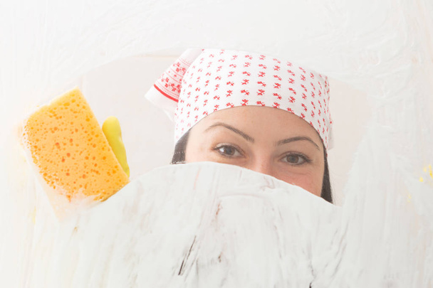 Female with bandana cleaning shower glass covered with foam with orange sponge - Φωτογραφία, εικόνα
