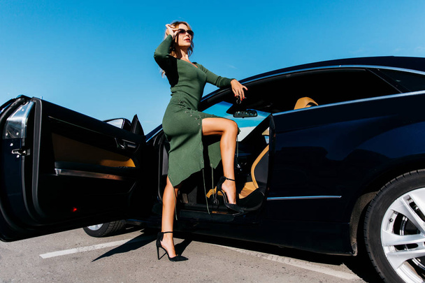 Photo of sexy blonde with keys in sunglasses in long dress standing near black car with open door - Zdjęcie, obraz