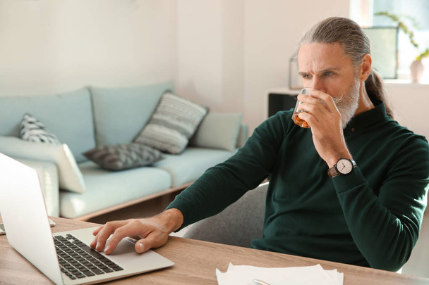 Hombre mayor bebiendo whisky mientras trabaja en el portátil en casa
 - Foto, imagen