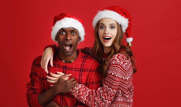 happy cheerful couple black man and caucasian woman in christmas hats on red backgroun - Φωτογραφία, εικόνα