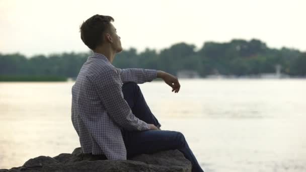Melancholic male teen sitting alone on stone near river and thinking about life - Záběry, video
