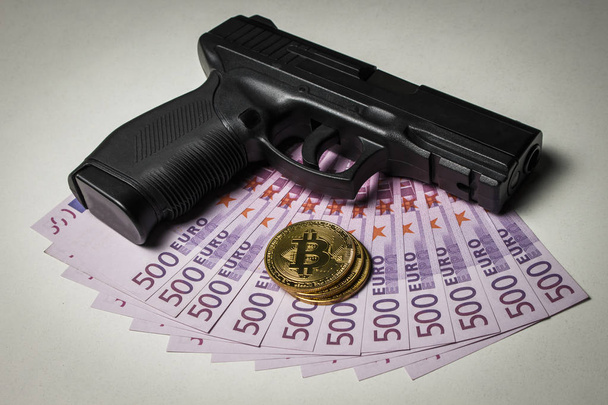 Pistola negra con un paquete de euros y monedas en forma de bitcoin sobre una mesa blanca
 - Foto, Imagen