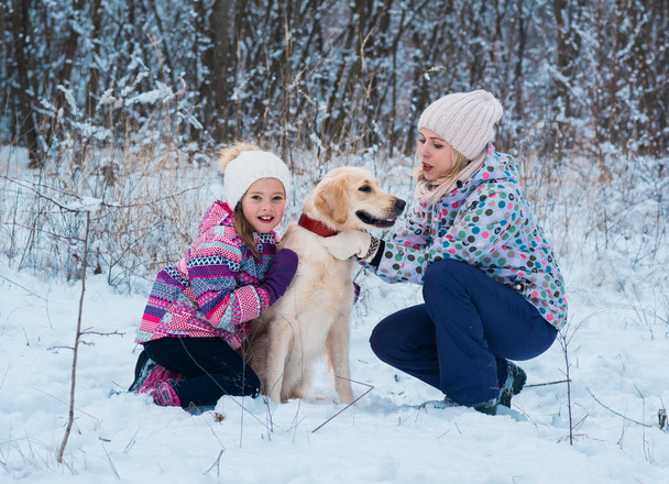 Feliz hermosa familia joven con su perro golden retriever en un día de invierno. Mujer e hija abrazándose con el perro en el campo nevado invierno. Amistad, mascotas y humanos
. - Foto, Imagen
