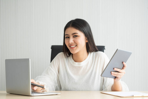 Ασιατικές γυναίκα κάνει freelance εργασία της με φορητό υπολογιστή και ψηφιακό tablet στο γραφείο στο σπίτι της. Χαμογελαστά γυναικεία βλέπουν φωτογραφική μηχανή. - Φωτογραφία, εικόνα