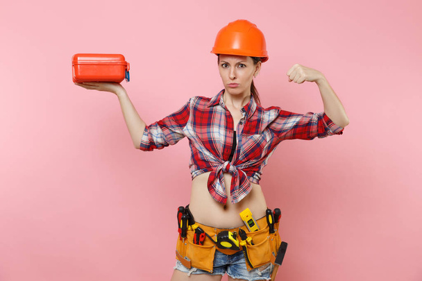Strong young woman in orange helmet, plaid shirt, denim shorts, kit tools belt full of instruments, toolbox showing biceps, muscles isolated on pink background. Female male work. Renovation concept - Foto, imagen