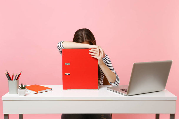 Donna stanca appoggiata alla cartella rossa con documenti cartacei e che lavora al progetto mentre è seduta in ufficio con laptop isolato su sfondo rosa pastello. Concetto di carriera aziendale di successo. Copia spazio
 - Foto, immagini