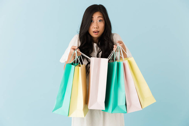 Image of asian young shocked emotional woman isolated over blue background holding shopping bags. - 写真・画像