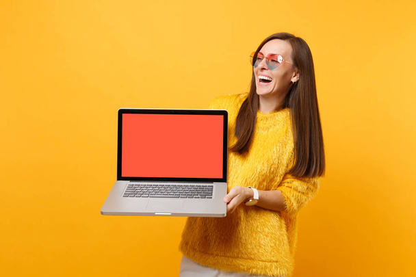 Rire jeune femme dans les lunettes de coeur tenant ordinateur portable PC ordinateur avec écran vide noir vierge isolé sur fond jaune vif. Les gens émotions sincères, concept de style de vie. Espace publicitaire
 - Photo, image