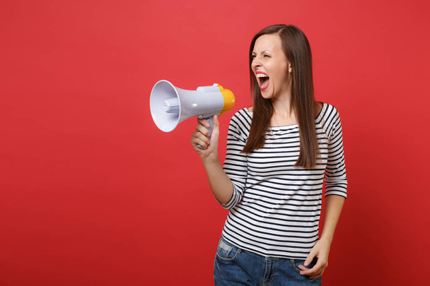 Portrait of irritated angry young woman in striped clothes looking aside, screaming on megaphone isolated on bright red wall background. People sincere emotions, lifestyle concept. Mock up copy space - Photo, Image