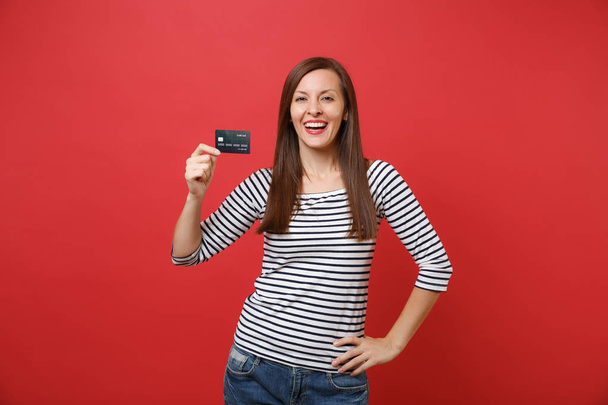 Portrait of smiling joyful young woman in casual striped clothes standing, holding credit card isolated on bright red wall background. People sincere emotions, lifestyle concept. Mock up copy space - Valokuva, kuva