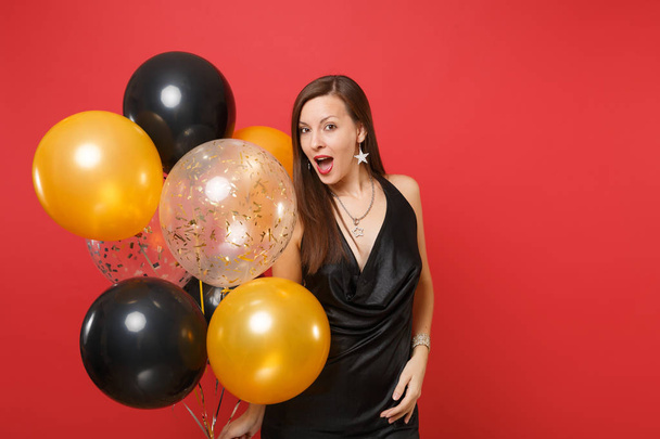 Amazed young woman in little black dress celebrating holding air balloons isolated on red background. St. Valentine's, International Women's Day, Happy New Year, birthday mockup holiday party concept - Photo, Image