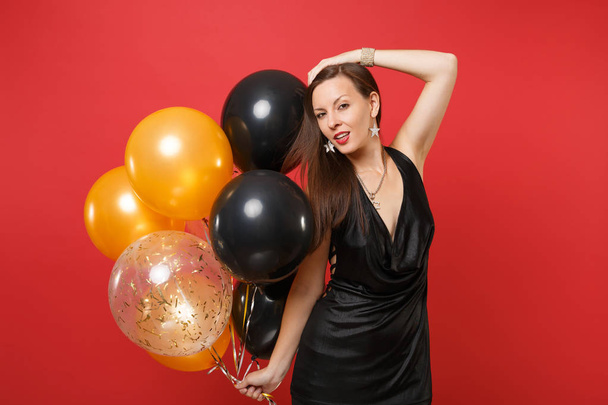 Chica joven pasión en pequeño vestido negro celebrando poner la mano en la cabeza mantenga globos de aire aislados en el fondo rojo. Día Internacional de la Mujer, Feliz Año Nuevo concepto de fiesta simulada de cumpleaños
 - Foto, imagen