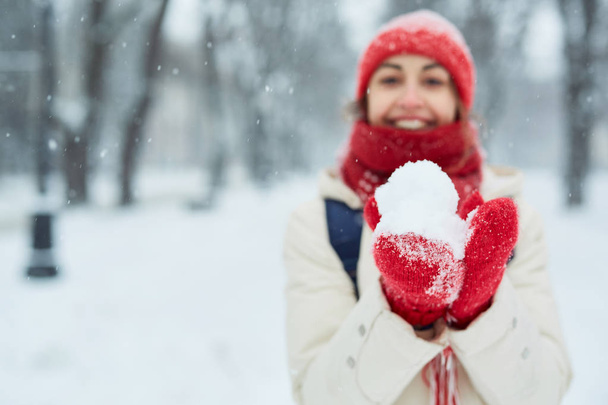 Cheerful smiling woman in white down jacket and red cap, scarf and mittens walking and plaing snowballs on the snowy street after blizzard in city. winter city after blizzards and snowfall. - Valokuva, kuva