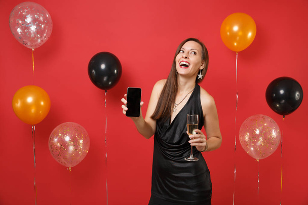グラス シャンパン、明るい赤の背景の気球に空白の黒い空の画面で携帯電話を保持している黒のドレスで陽気な若い女性。幸せな新年、誕生日モックアップ休日党概念 - 写真・画像