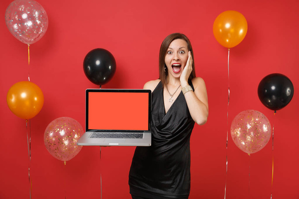 Fille choquée en robe noire gardant la main sur le visage tenant ordinateur portable PC ordinateur avec écran vide noir vierge sur des ballons d'air de fond rouge vif. Bonne année, concept de fête de fête anniversaire maquette
 - Photo, image