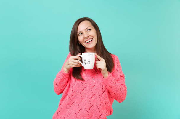 Smiling young woman in knitted pink sweater looking up and hold in hands cup of coffee or tea isolated on blue turquoise wall background, studio portrait. People lifestyle concept. Mock up copy space - Photo, Image