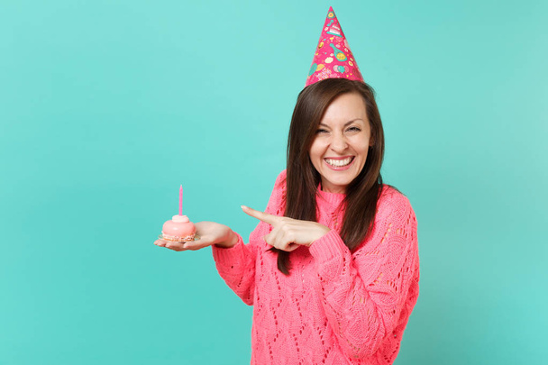 Lustige junge Frau in gestricktem rosa Pullover Geburtstagsmütze zeigt Zeigefinger auf Kuchen mit Kerze in der Hand isoliert auf blauem Wand Hintergrund Studio-Porträt. Menschen Lebensstil Konzept. Attrappe Kopierraum - Foto, Bild