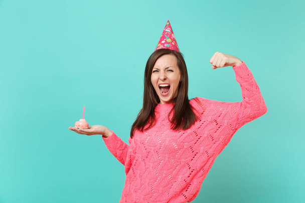Crazy young woman in knitted pink sweater, birthday hat screaming, showing biceps, muscles holding in hand cake with candle isolated on blue background. People lifestyle concept. Mock up copy space - Photo, Image