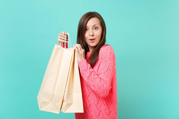 Amazed beautiful young woman in knitted pink sweater hold in hands packages bags with purchases after shopping isolated on blue turquoise wall background. People lifestyle concept. Mock up copy space - Photo, Image