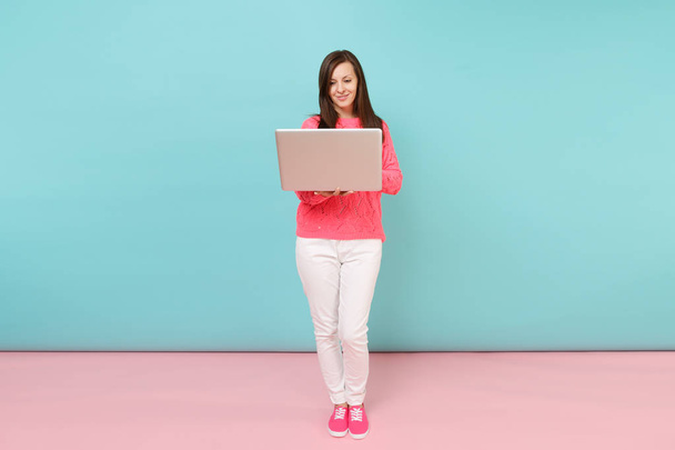 Full length portrait of woman in knitted sweater, white pants using laptop pc computer isolated on bright pink blue pastel wall background in studio. Fashion lifestyle concept. Mock up copy space - Photo, Image
