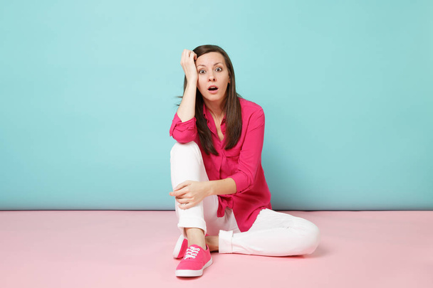 Retrato de larga duración de una joven conmocionada en blusa de camisa rosa, pantalones blancos sentados en el suelo aislados en un estudio de fondo de pared pastel azul rosado brillante. Concepto de estilo de vida de moda Simular espacio de copia
 - Foto, imagen