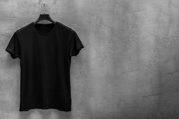 Front side of male black cotton t-shirt on a hanger and a concrete wall in the background. T-shirt without print and copyspace for your text on right side - Fotó, kép