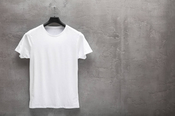 Front side of male white cotton t-shirt on a hanger and a concrete wall in the background. T-shirt without print and copyspace for your text on right side - Foto, Imagem