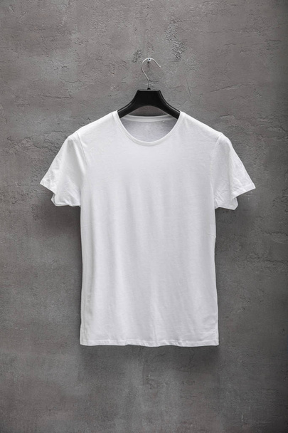 Front side of male white cotton t-shirt on a hanger and a concrete wall in the background. T-shirt without print - Foto, immagini